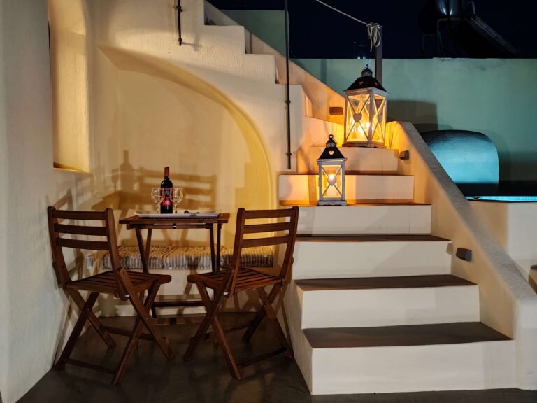 A small outdoor wooden table with two chairs, a wine bottle, and a glass beside lit lanterns on steps against a curved, cream-colored wall at night.